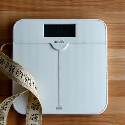 A sleek, minimalist digital scale paired with measuring tape and a notebook, suggesting precision in tracking body composition. Clean, clinical aesthetic with subtle reflections. Prominent colors: background light, primary light, and text secondary.